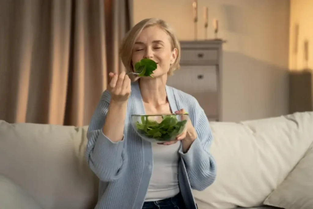 Evde koltukta otururken cam kaseden yeşil salata yiyen kadın